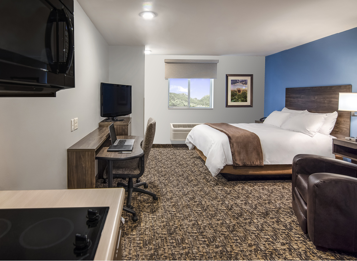 Interior hotel room facing window, with kitchenette, rolling desk and office chair, TV, single queen bed and brown recliner with a blue accent wall.