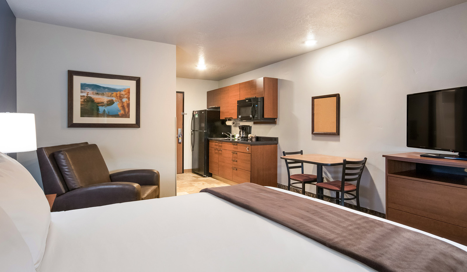 Interior hotel room facing the door with a single queen bed, brown recliner, dresser and TV, table and chairs, and kitchenette.