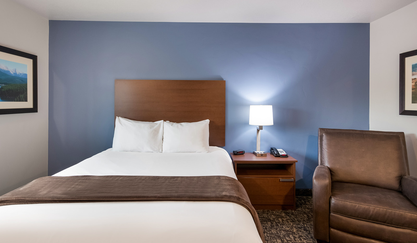 Interior hotel room with single queen bed, bedside table and lamp, brown recliner and blue accent wall.