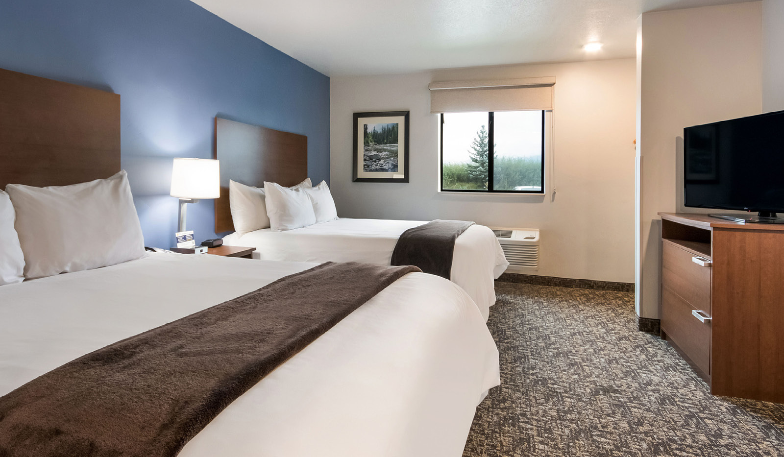 Interior hotel room with two queen beds against a blue accent wall across from a dresser with TV on top.