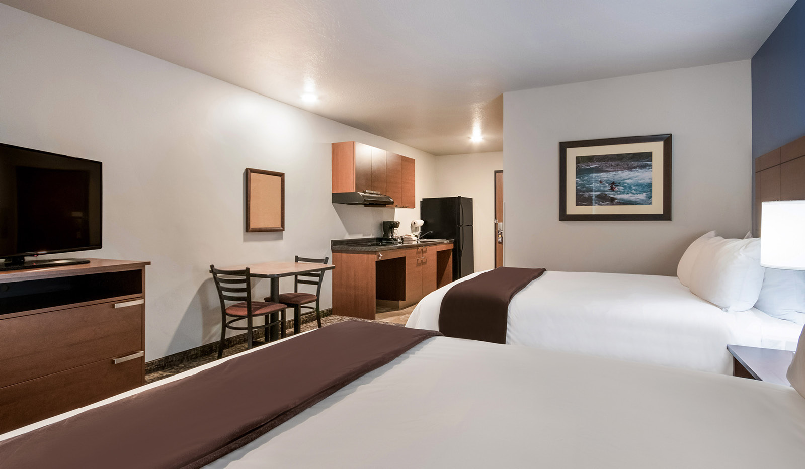 Interior hotel room facing door with two queen beds across from a dresser with a TV on top, table and chairs, and kitchenette.