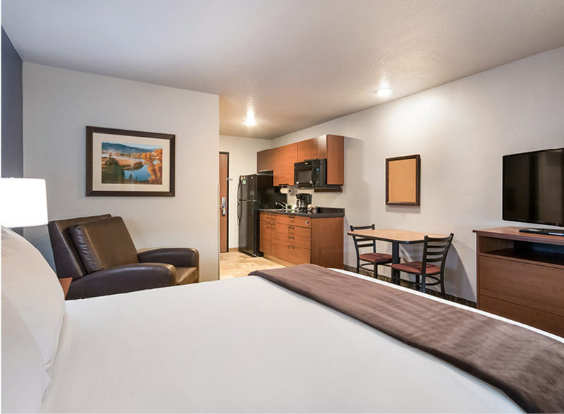 Interior hotel room facing the door with single queen bed, brown recliner, kitchenette, table and chairs, and dresser with a TV on top.