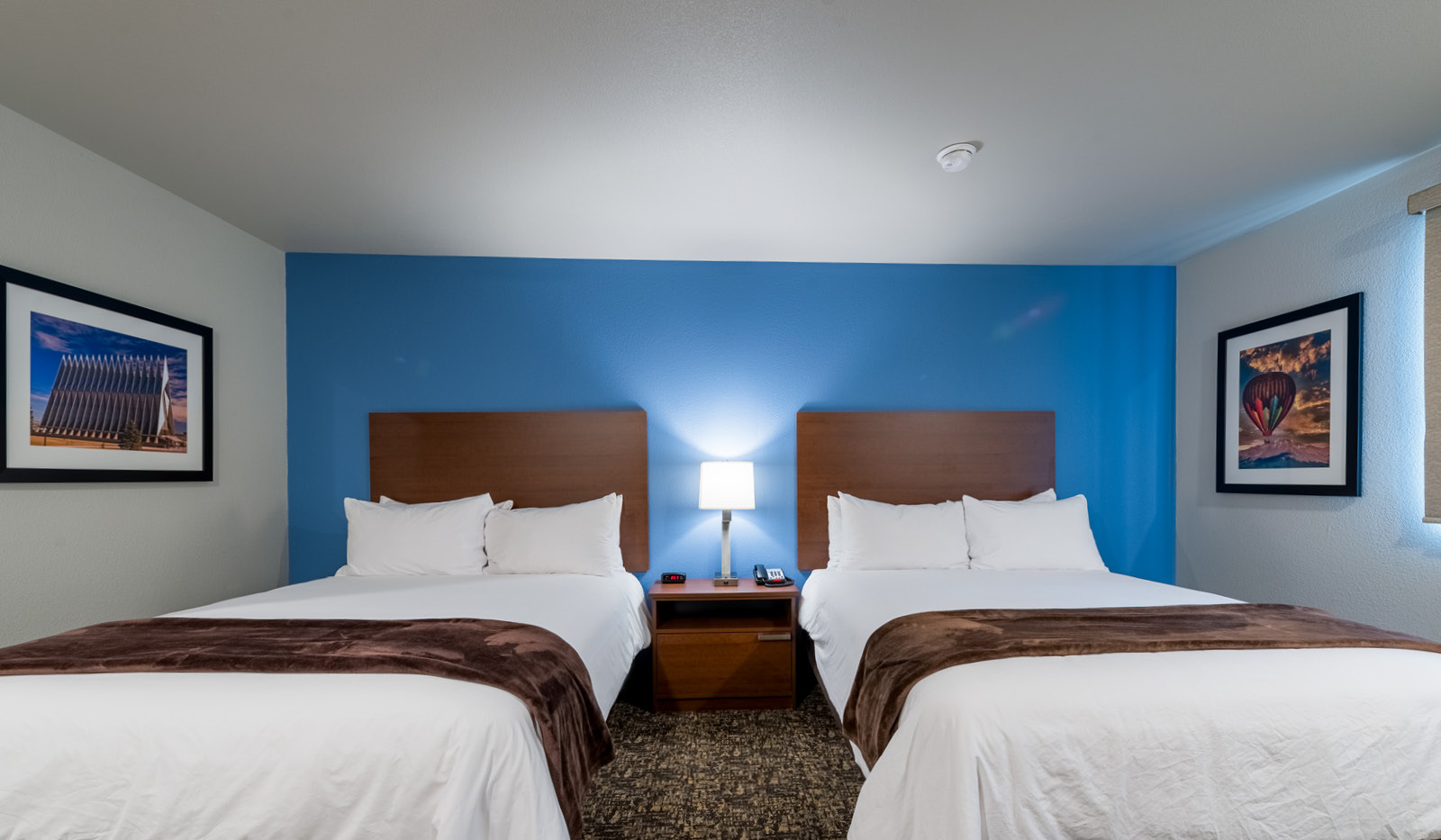 Interior hotel room with bedside table and lamp flanked by two queen beds and a blue accent wall.