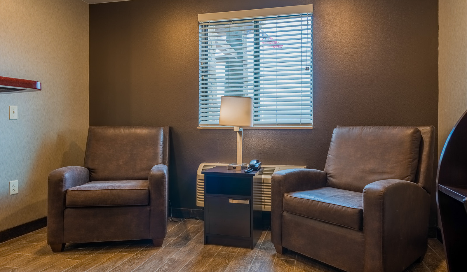 Interior lobby with small table and lamp flanked by two brown recliners, setting in front of a brown accent wall.