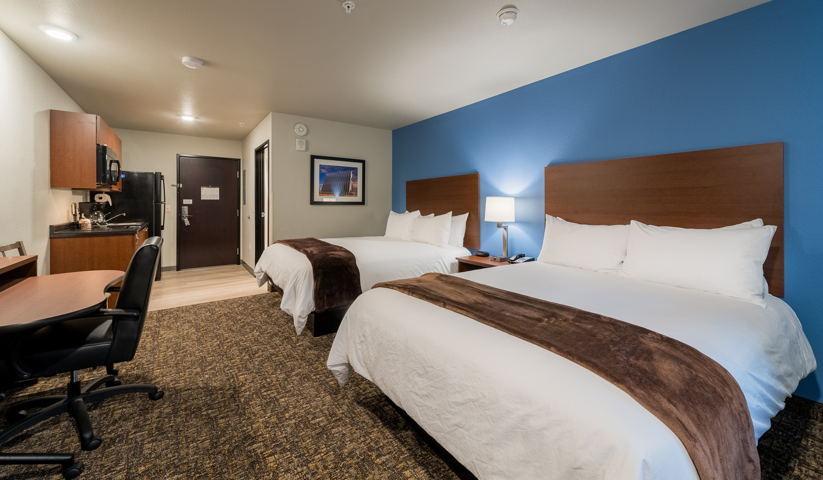 Interior hotel room facing the door, with two queen beds, rolling desk and chair, kitchenette. and blue accent wall.