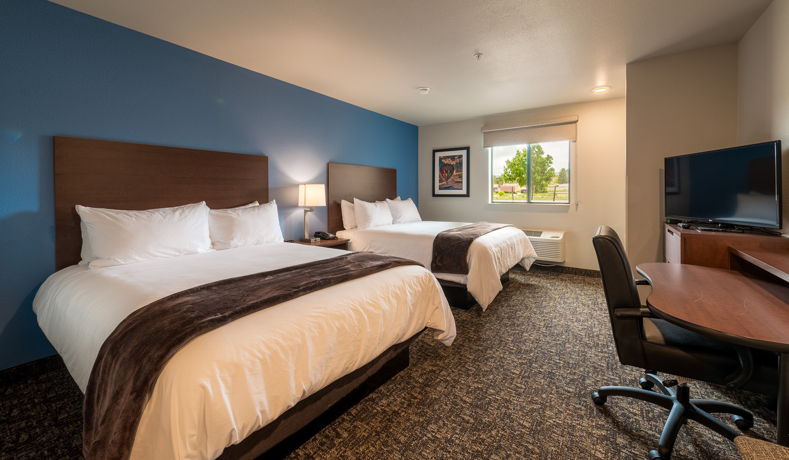 Interior hotel room with two queen beds, dresser, television, desk, chair, and blue accent wall.