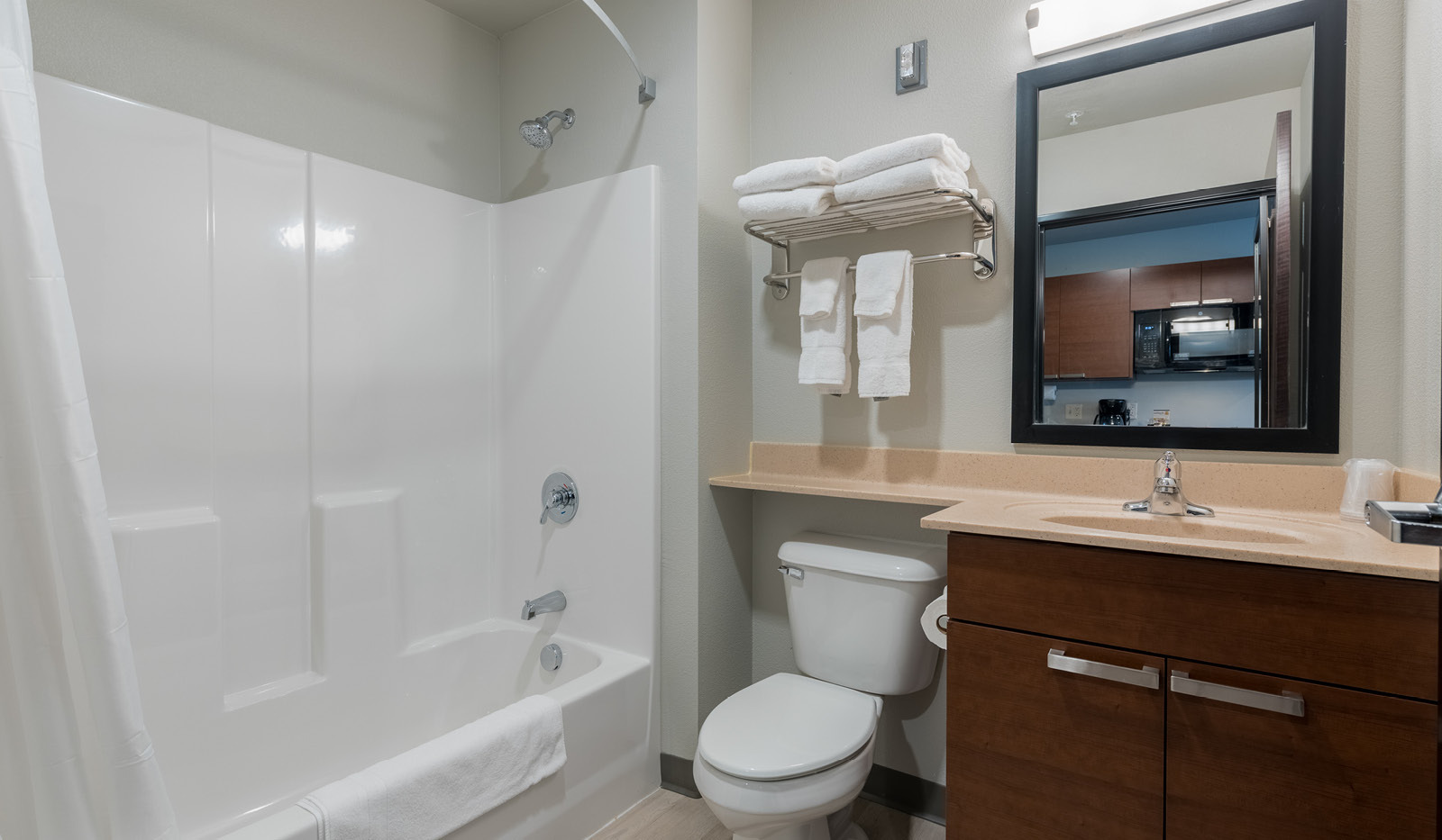 Interior bathroom with white shower and tub, folded towels in a rack above a toilet, with vanity and mirror on the right.