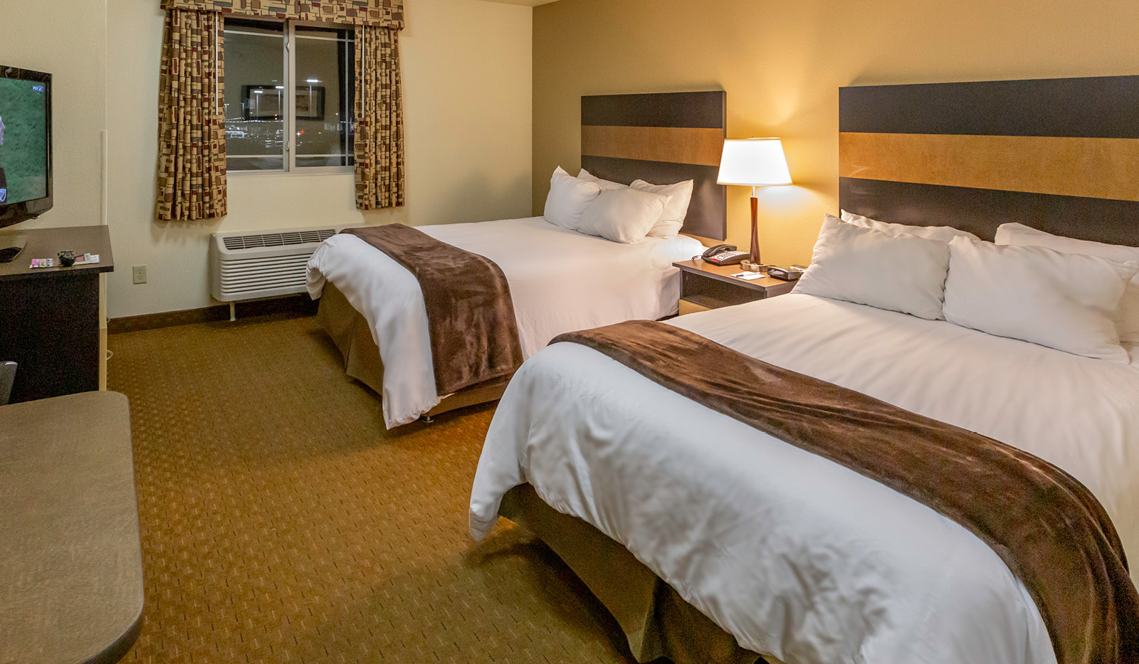 Interior hotel room with two queen beds, table and lamp, table and chairs, dresser and TV.