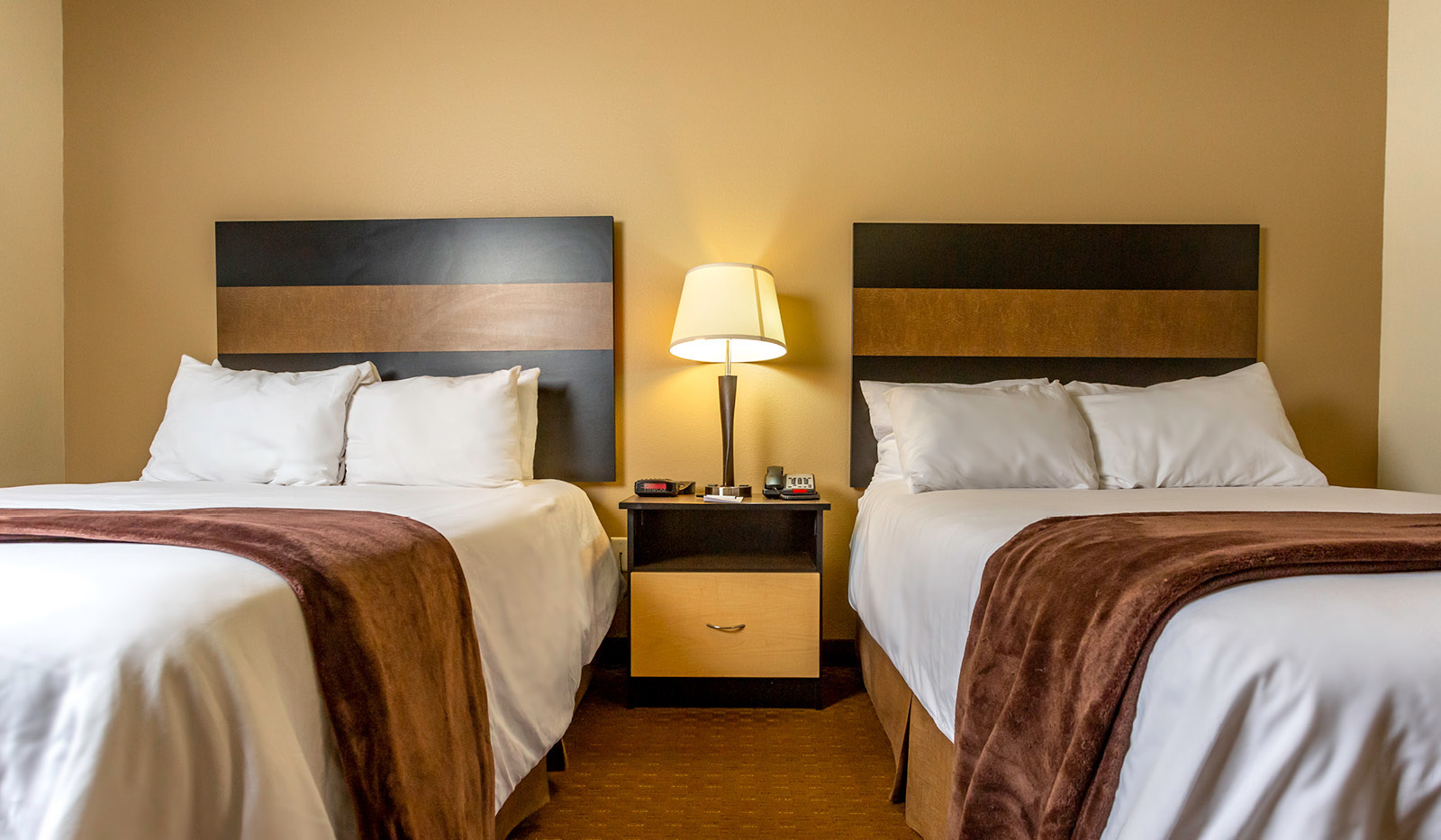 Interior hotel room with bedside table and lamp flanked by two queen beds and a tan accent wall.