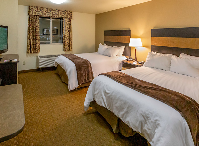 Interior hotel room with two queen beds, table and chairs, dresser, TV and tan accent wall.