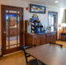 Hotel breakfast area with an updated self-serve coffee station on a wooden cabinet, surrounded by tables, glass-paneled doors, and warm interior lighting.
