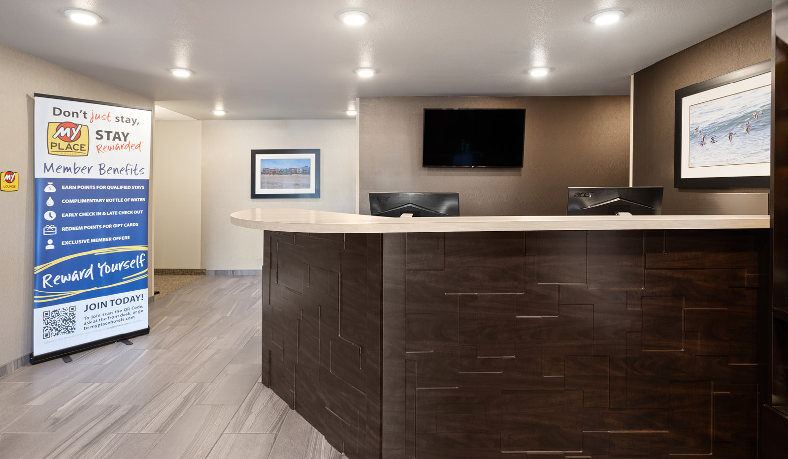 Wooden front desk with tan countertop in front with lobby banner, tv and photos surrounding.