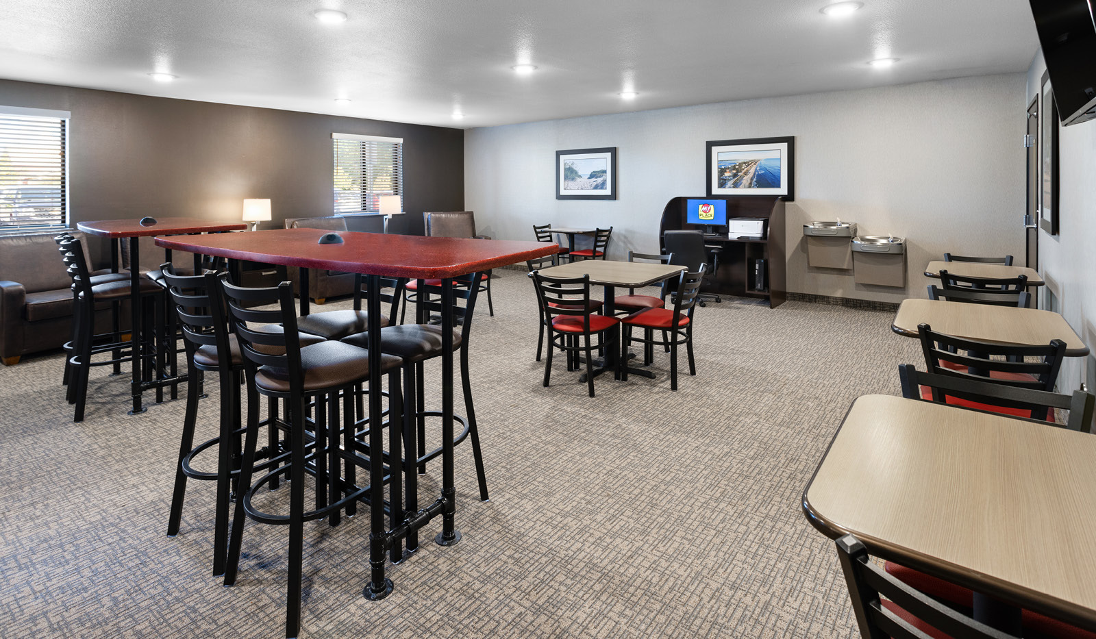 Multiple rows of high-top and regular height tables in the lobby lounge with the computer and printer in the back next to a drinking fountain.