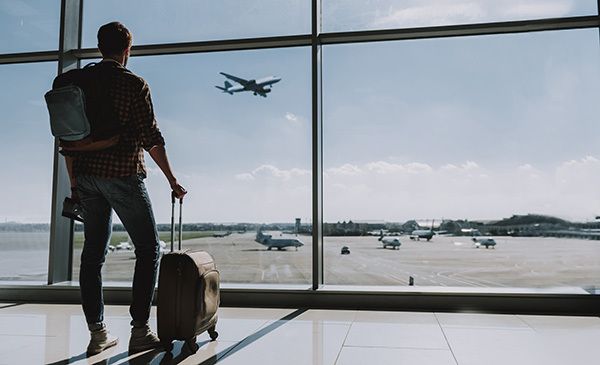 A man with backpack and luggage staring out the window at the airport.