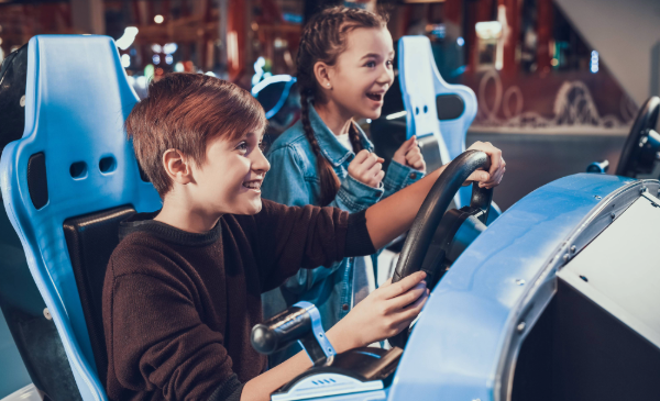 A young boy sits in a blue gaming console holding a steering wheel while a young girl with braids cheers him on next to him.