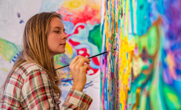 A side view of a woman with long blonde hair uses a black paintbrush to paint a colorful mural.