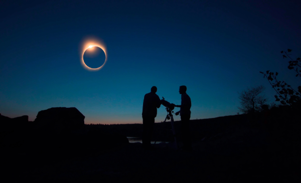 The silhouette of two people around a telescope under a dark blue sky, looking at an eclipse.