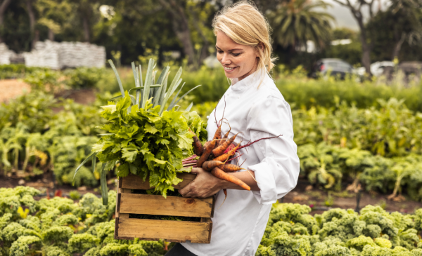 A blond woman, wearing a white chefs coat, stands in a large vegetable garden while she holds a wooden box filled with carrots, lettuce, and other vegetables.