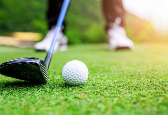 Closeup of a golf ball and a club next to it, with a person's feet in the background.