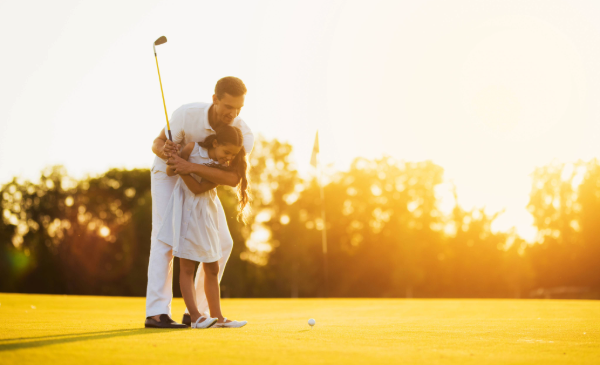 A man stands behind a young girl on a golf course as he shows her how to swing a golf club.