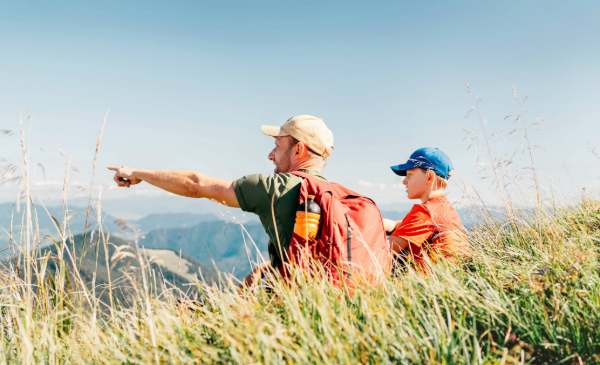 A man points into the distance while a child looks on, seated in a grassy area overlooking a mountain backdrop.