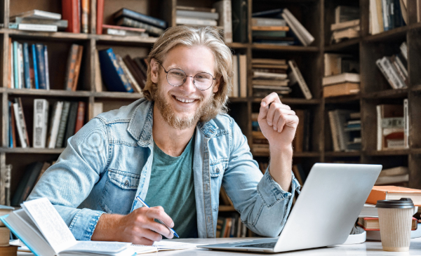 A man smiles at the camera while working on a computer surrounded by books.