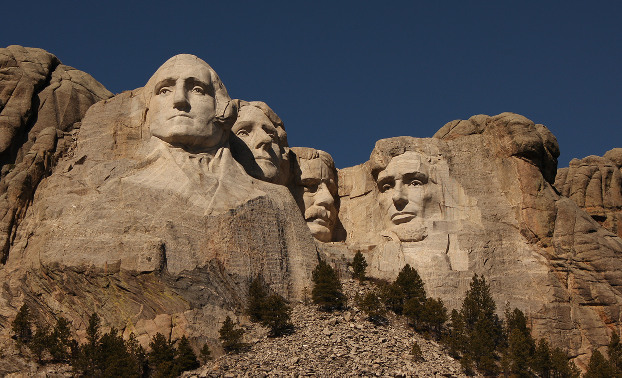 Four presidential faces carved into the side of a mountain with large evergreen trees below.