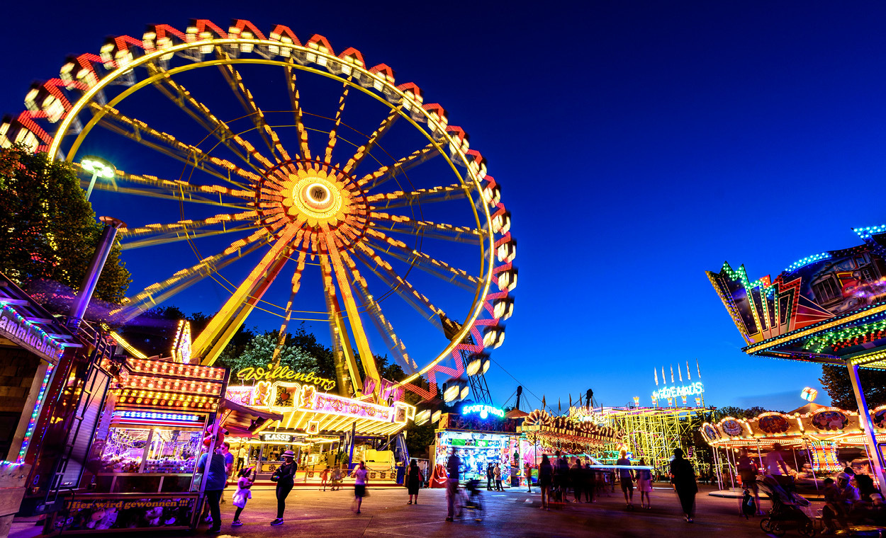 Ferris wheel lit up behind small neon booths under a night sky.