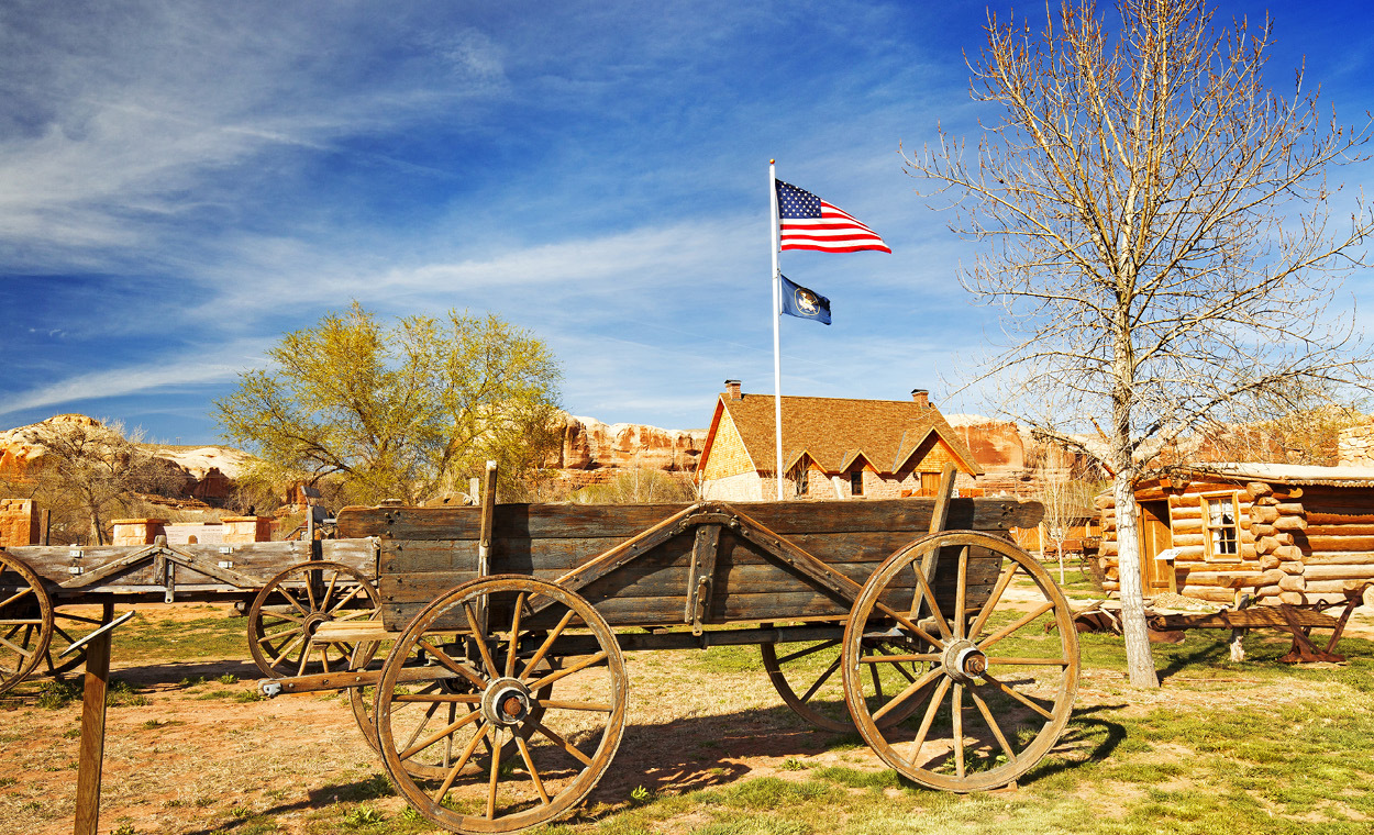 Wooden wagon with wooden wheels in front of a United States flag and multiple historic buildings.