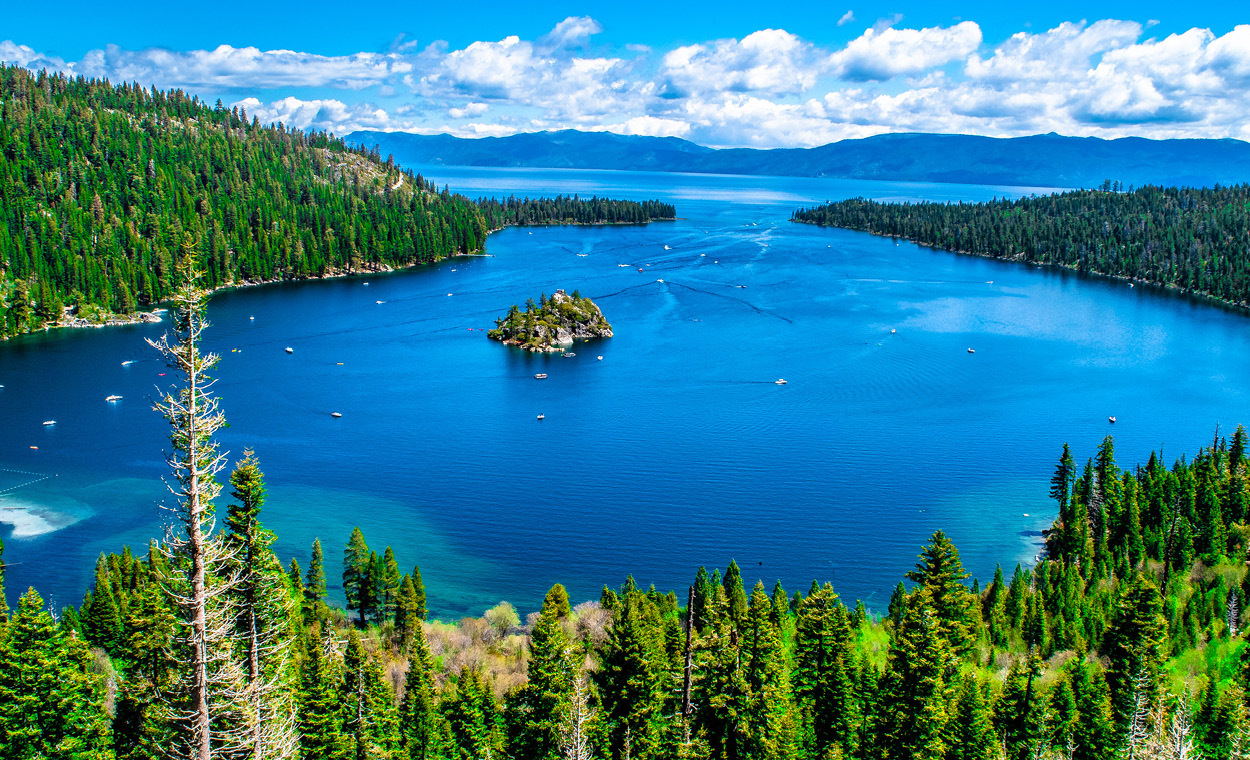 Large blue lake surrounded by pine trees with mountains in the distance.