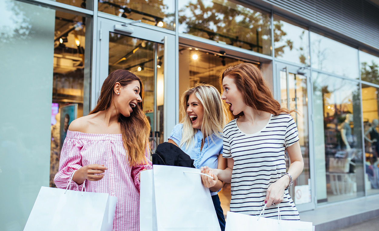 Three women talking while holding shopping bags outside a clothing store.