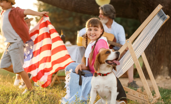 A young girl sits in a lawn chair while petting a small brown and white dog, while a boy runs with an American flag in the background.
