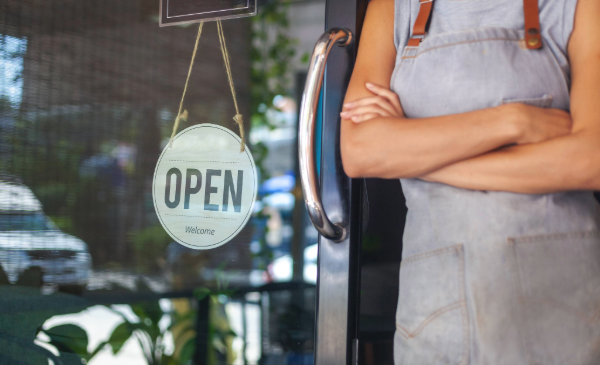 A woman wearing a gray apron stands with her arms folded next to a glass door with an "Open" sign hanging.