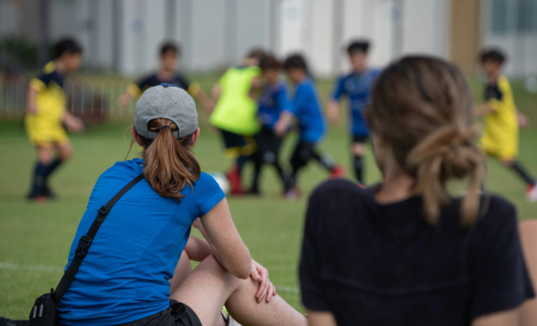 Two women sit and watch children playing soccer.