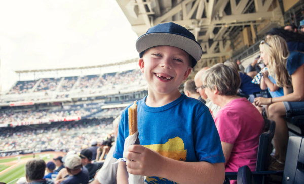 A young boy smiles at the camera while eating a snack at a baseball game.