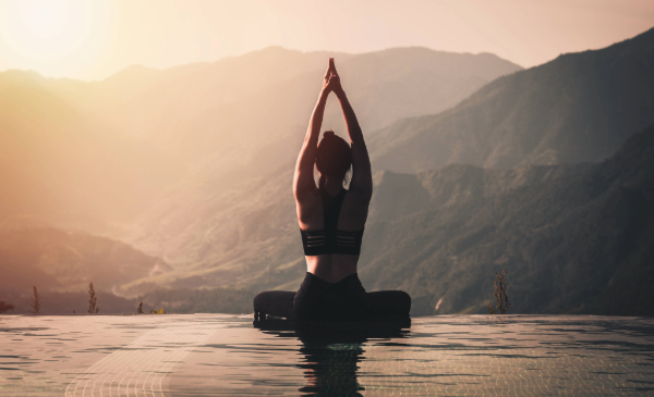 A woman reaches closed hands towards the sky as she sits in a yoga pose before a body of water.