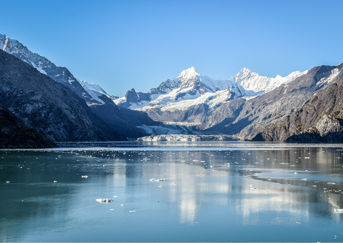 A clam lake sets before a large, snowcapped mountain range under a clear blue sky.