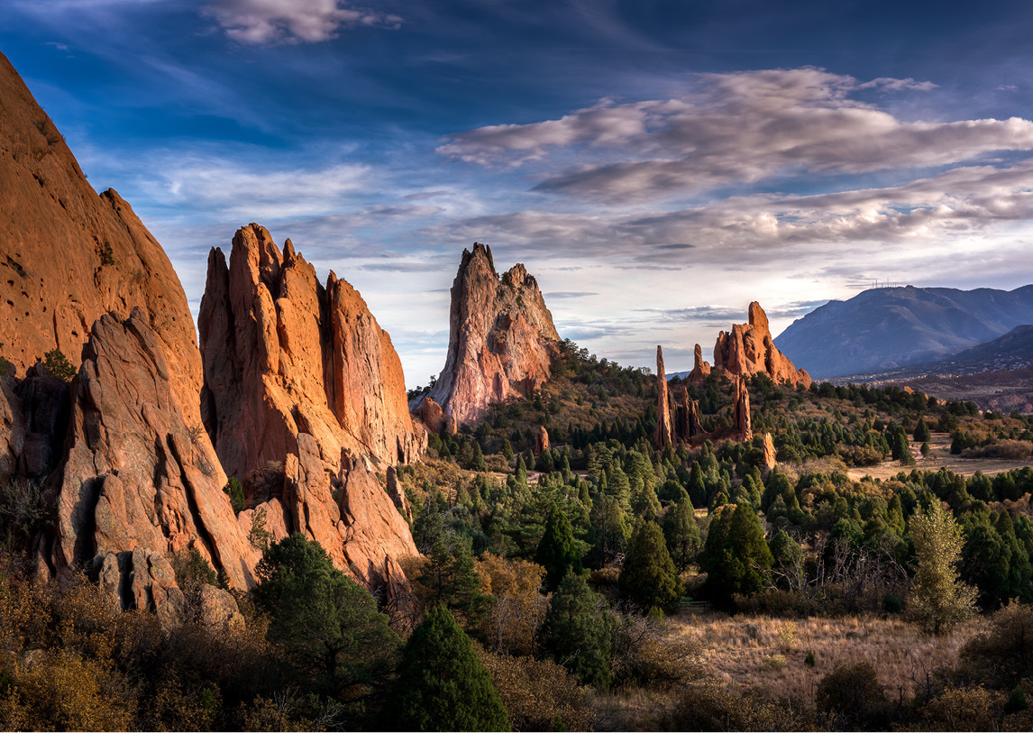 Natural rock formations surround a valley of green trees under a blue, cloudy sky.