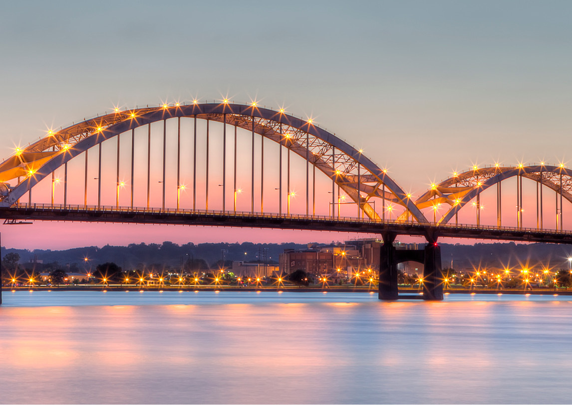 An arched, lit bridge sits above a calm river with lighted buildings in the background.
