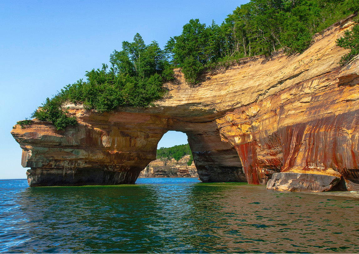 A body of water faces a large rock formation covered in green trees with a natural stone arch over the water.