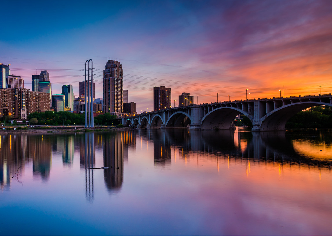 A bridge sets above calm water as it reflects and connects with a populous downtown with tall buildings, all under a blue and orange sunset.