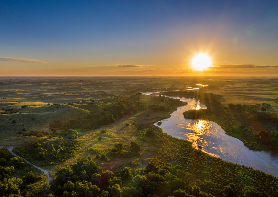 A calm river winds between green grasses and trees with a setting sun in the background.