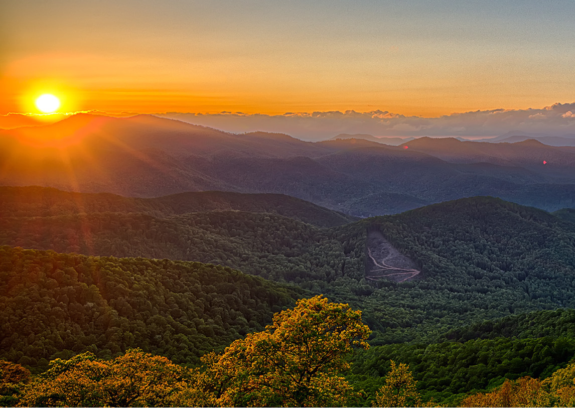 A green valley sets before a mountainous background with an orange sunset.