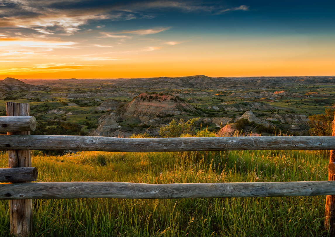 A wooden fence sets before a wide valley of natural rock formations and prairie grasses under a wide sunset.