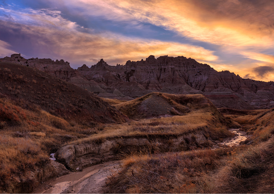 Natural rock formations called The Badlands set behind prairie grasses under a blue and orange sunset.