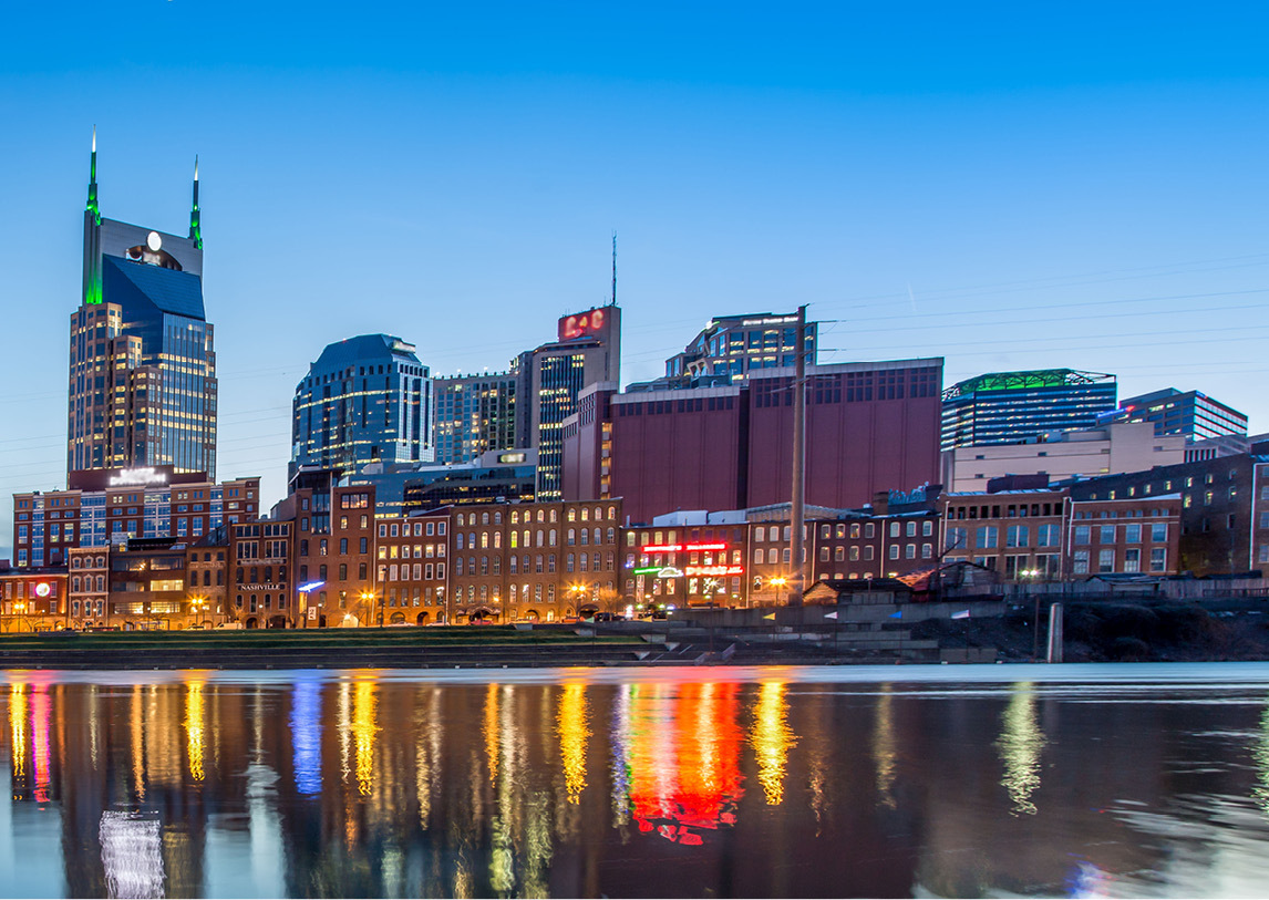 Profile of a city skyline under clear blue skies, with the cityscape reflecting off a body of water in the foreground.
