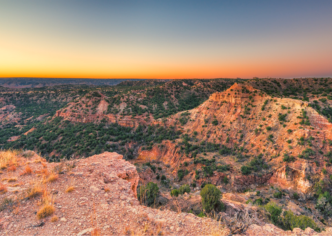 An orange rock cliff overlooks large natural rock formations and a forest of green trees.