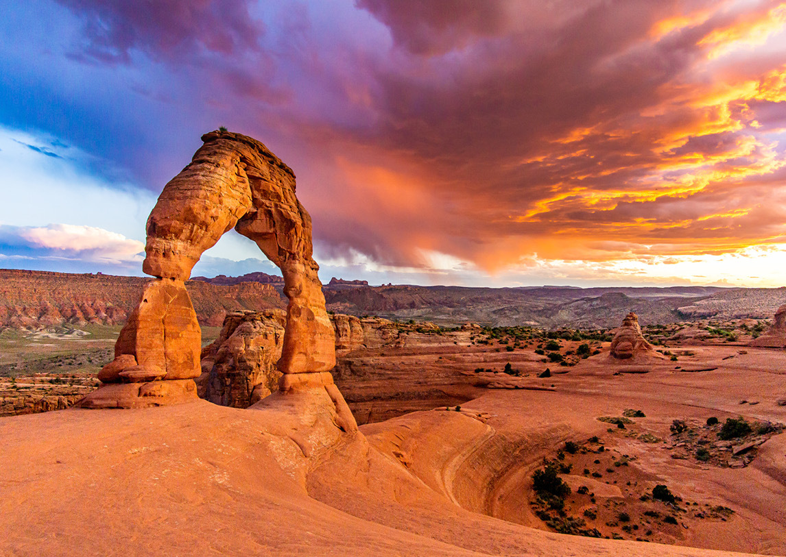 An arched rock formation sits before a vast layout of orange rocks at Arches National Park, under a blue, purple and pink sunset.