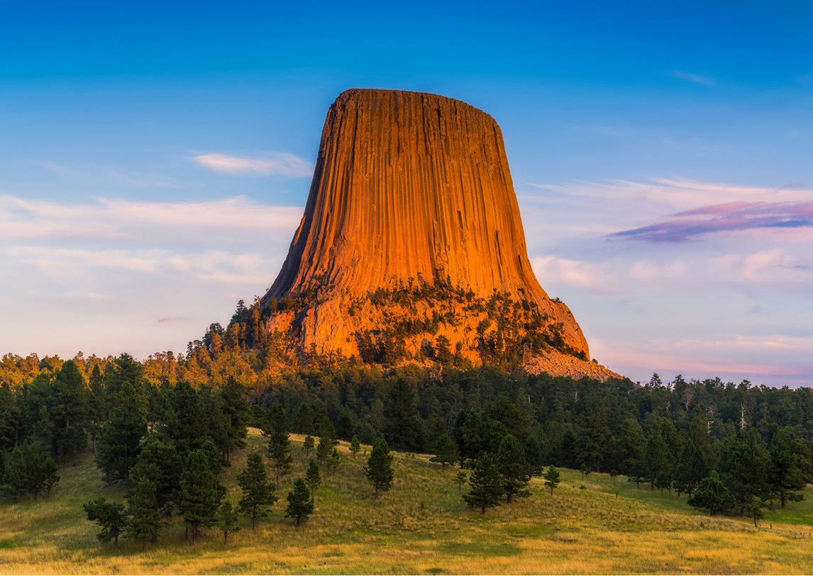 A tall, orange plateau called Devil's Tower rises above a group of trees under partly cloudy skies.
