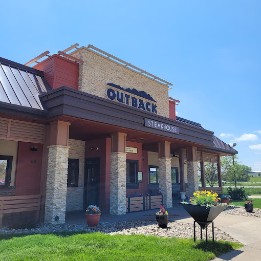 Exterior view of a red and brick building. The entrance has multiple columns and numerous flower pots. The sign above the door says "Outback steak" in blue lettering.